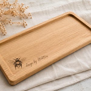 Può includere: Un vassoio rettangolare in legno marrone chiaro con centro incassato e angoli arrotondati. La superficie presenta un'illustrazione incisa al laser di un bombo seguita dal testo in corsivo design by Wehtras nell'angolo in basso a sinistra.