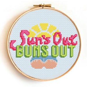 May include: A cross-stitch embroidery hoop with a white background and a colourful design that says "Sun's Out Buns Out" with a yellow sun and two peach-coloured buns.