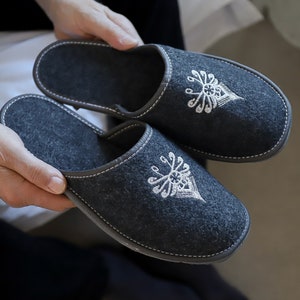 Puede incluir: Un par de zapatillas de fieltro gris con diseños bordados en blanco. Las zapatillas tienen un ribete blanco cosido alrededor de los bordes.