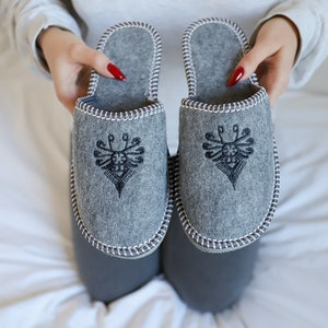 Puede incluir: Un par de zapatillas de fieltro gris con diseños bordados en negro. Las zapatillas tienen un ribete blanco alrededor de los bordes.