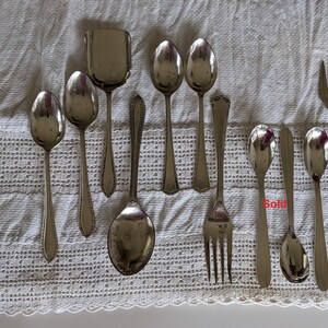 Puede incluir: Una colección de cubiertos vintage, que incluye cucharas, un tenedor y una cuchara para servir. Los cubiertos están dispuestos sobre un mantel de crochet blanco.