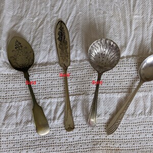 Könnte beinhalten: Vier antike Servierutensilien aus Silber, darunter ein Löffel mit aufwendigen floralen Details, ein Buttermesser, ein muschelförmiger Löffel und ein einfacher Löffel. Die Utensilien werden auf einer weißen, strukturierten Oberfläche präsentiert.