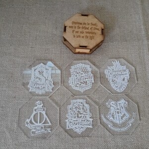 May include: Set of six clear glass coasters with white etched designs of the Hogwarts house crests. The coasters feature the crests for Gryffindor, Slytherin, Hufflepuff, Ravenclaw, and the Deathly Hallows symbol. The coasters are packaged in a wooden box with a quote about happiness.