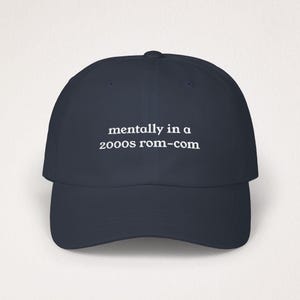 Puede incluir: Gorra de béisbol azul marino con el texto blanco "mentally in a 2000s rom-com". La gorra tiene una visera curva y una corona redonda. Un accesorio de moda para un look casual.