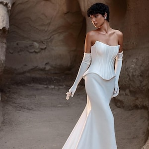 Puede incluir: Vestido de novia blanco sin tirantes con corpiño ajustado y falda fluida. El vestido presenta un top estilo corsé y guantes largos blancos. El vestido tiene una larga cola. La persona está en un entorno natural al aire libre.