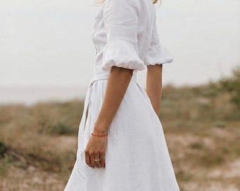 May include: A white linen dress with short, ruffled sleeves and a flowing skirt. The dress has a cinched waist and a wrap-style design. The model is standing on a wooden walkway near the beach.