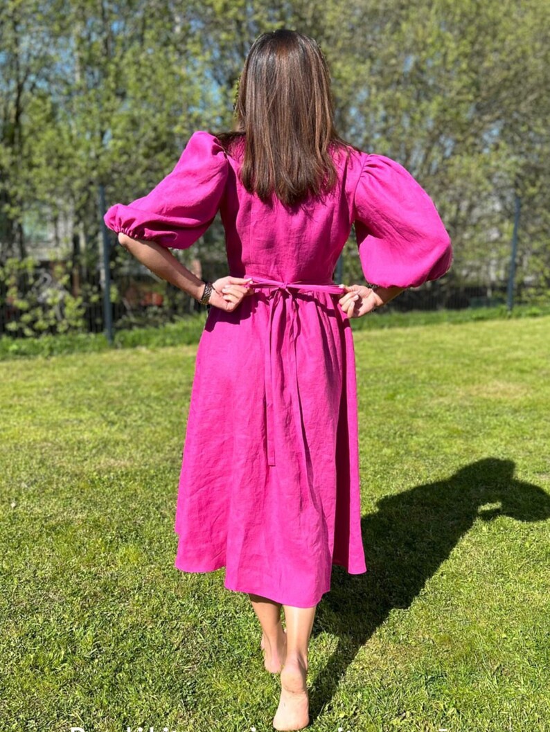 Puede incluir: Un vestido de lino rosa con cintura ce&ntilde;ida y mangas abullonadas. El vestido est&aacute; atado en la espalda con una cinta a juego.