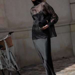 May include: A person in a black outfit, including a sheer top over a tank top and a long skirt, holds a black handbag. They wear sunglasses and earrings. A bicycle with a basket is in the background. The outfit is elegant and stylish.