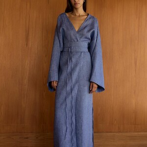 以下が含まれることがあります： Vネックとリボンベルトが付いた、長い青いリネンラップドレス。ドレスは長袖で、流れるようなシルエットです。