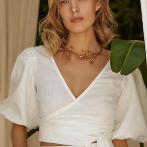 May include: A white linen wrap top with puffed sleeves and a gold floral choker necklace. The top is tied at the waist and the sleeves are three-quarter length.