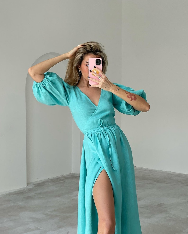 Puede incluir: Un vestido de lino turquesa con falda larga, cuello en V y un lazo en la cintura. El vestido tiene una abertura en la pierna izquierda.