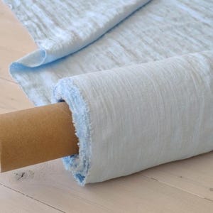 Puede incluir: Un rollo de tela azul claro con un borde sin rematar, parcialmente desenrollado para revelar la textura. La tela está enrollada alrededor de un tubo de cartón marrón. La tela es de color azul claro.