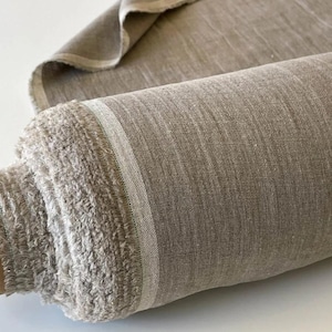 May include: A roll of natural-colored linen fabric. The fabric is a neutral beige-brown with a subtle texture and a light-colored stripe along the edge. The edges of the fabric are slightly frayed, giving it a rustic appearance. The fabric is rolled up.