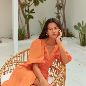 Puede incluir: Una mujer con un vestido de lino naranja brillante con mangas abullonadas se sienta en una silla de mimbre. El vestido tiene un escote en V y un estilo de envoltura. La mujer lleva pendientes de aro dorados y un anillo dorado.