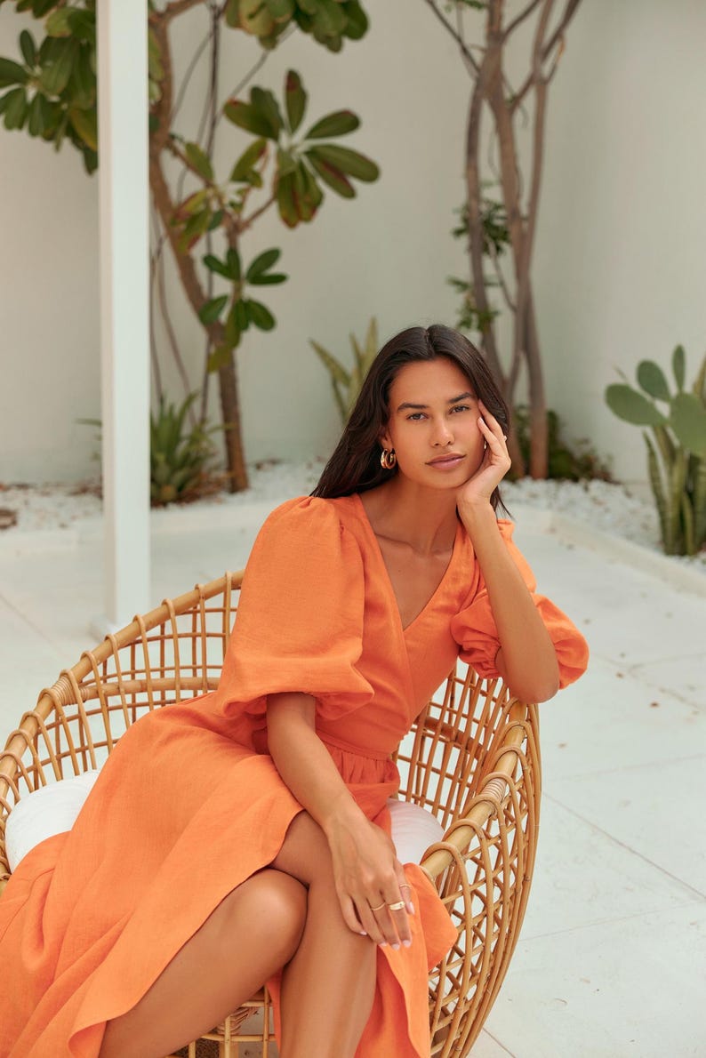 Puede incluir: Una mujer con un vestido de lino naranja brillante con mangas abullonadas se sienta en una silla de mimbre. El vestido tiene un escote en V y un estilo de envoltura. La mujer lleva pendientes de aro dorados y un anillo dorado.