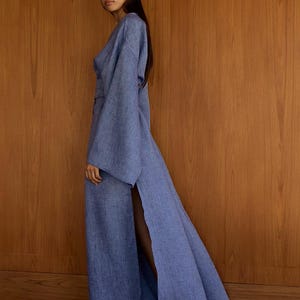 以下が含まれることがあります： ラップスタイルのボディスと長袖の薄いブルーのリネンロングドレス。ドレスはサイドスリットとウエストにベルトが付いています。