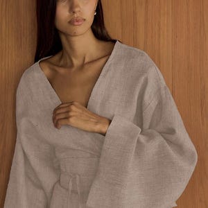 以下が含まれることがあります： 女性が薄いグレーのリネン素材のラップドレスを着ています。長袖でVネックです。ドレスはウエストにリボンが付いています。