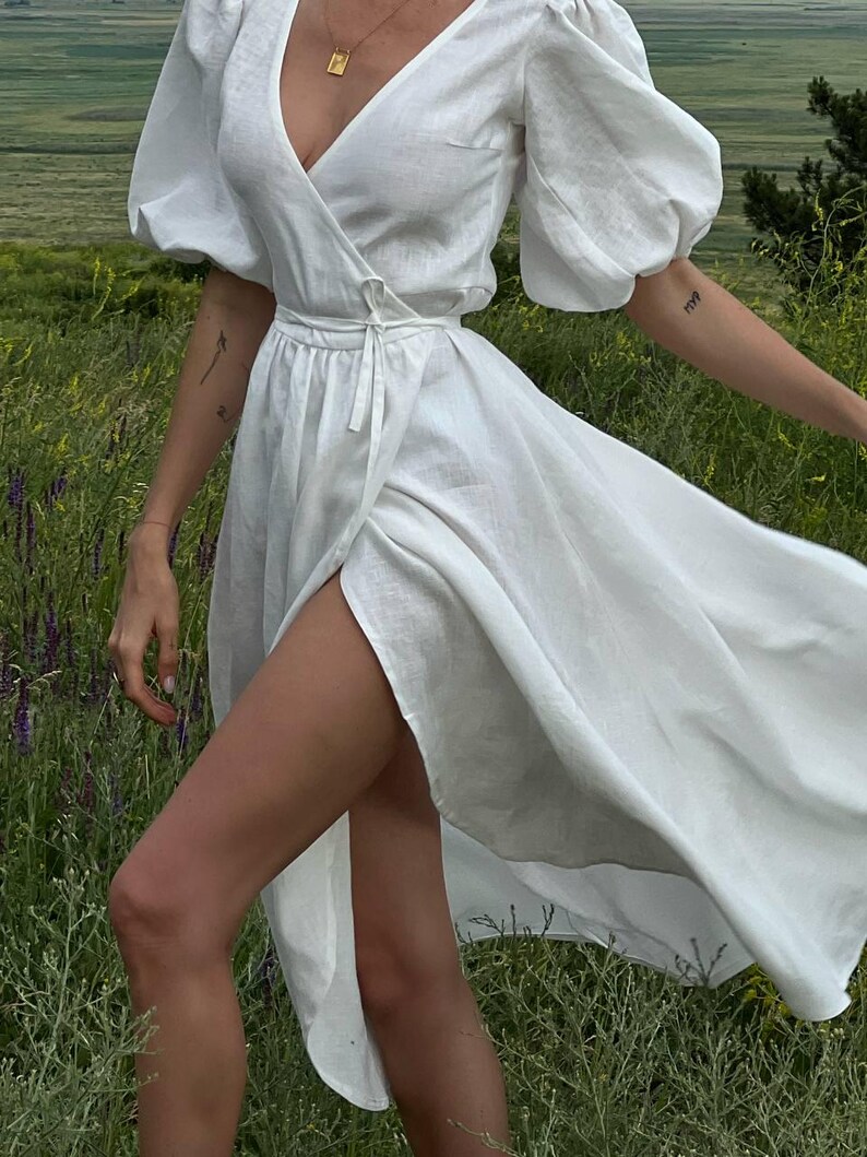 Puede incluir: Un vestido blanco de lino con cuello en V, mangas abullonadas y una abertura en el muslo. El vestido est&aacute; atado a la cintura con una cinta a juego.