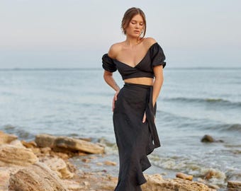 May include: A black two-piece outfit, consisting of a crop top with short sleeves and a wrap skirt. The skirt has a ruffled hem. The outfit is worn with black sandals. The background features a beach with rocks and the ocean.