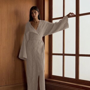 以下が含まれることがあります： Vネックと長袖の薄い茶色のリネン素材のロングドレスを着た女性。ドレスはウエストにリボンベルトが付いており、フロントにスリットが入っています。彼女は木製の壁と窓のある部屋に立っています。
