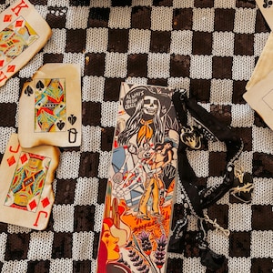 May include: A colorful, illustrated cigarette case with the text "ALWAYS BEEN CRAZY" and a black ribbon with heart charms. Playing cards and a leopard print bag are also visible on a black and white checkered surface.