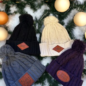 May include: Four knitted winter hats with pom-poms. The hats are in black, cream, grey, and purple. Each hat has a leather patch with a different saying. The sayings are "Perfectly Imperfect", "Perfectly Imperfect", "Faith + Fear Lead", and "Fearfully and Wonderfully Made".