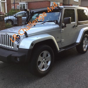 Könnte beinhalten: Ein silberner Jeep Wrangler 4x4 mit schwarzen Akzenten und einem schwarzen Dach. Der Jeep hat eine schwarze Stoßstange, schwarze Trittbretter und schwarze Räder. Der Jeep ist auf einer gepflasterten Auffahrt geparkt.