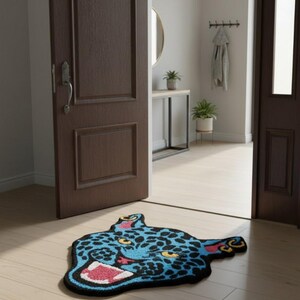May include: A blue and black rug shaped like a jaguar head with a wide open mouth. The rug has yellow spots, pink inner mouth, and red accents. The rug is placed on a light wood floor in front of a brown door.