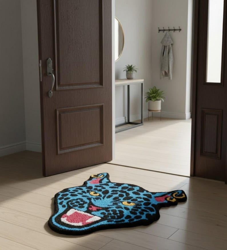 May include: A blue and black rug shaped like a jaguar head with a wide open mouth. The rug has yellow spots, pink inner mouth, and red accents. The rug is placed on a light wood floor in front of a brown door.
