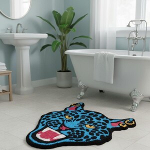 May include: A blue and black rug in the shape of a jaguar's head with yellow eyes and a pink mouth. The rug is on a tiled floor in a bathroom setting, with a white bathtub and sink visible in the background.