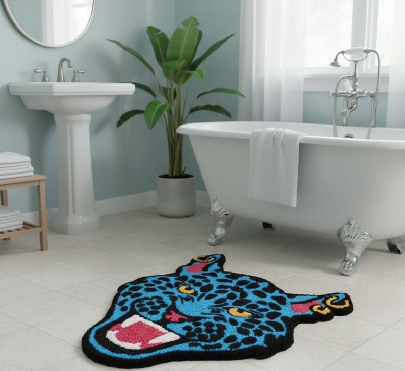 May include: A blue and black rug in the shape of a jaguar's head with yellow eyes and a pink mouth. The rug is on a tiled floor in a bathroom setting, with a white bathtub and sink visible in the background.