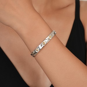 May include: A silver-toned cuff bracelet with a hammered, textured surface. The bracelet features three small, gold-toned star-shaped accents spaced evenly along the centre of the band, creating a contrast between the silver and gold metal tones.