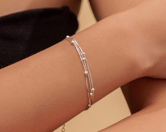 Può includere: Un delicato braccialetto d'argento con tre fili e piccole perle d'argento distanziate uniformemente. Il braccialetto è mostrato su un polso, con una semplice chiusura visibile. Il gioiello è di design minimalista.