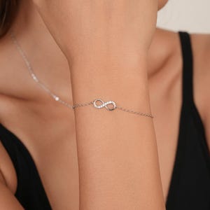 Könnte beinhalten: Ein zartes silberfarbenes Kettenarmband mit einem Unendlichkeitssymbol als zentralem Element. Das Unendlichkeitssymbol ist mit kleinen, klaren, funkelnden Steinen besetzt. Das Armband besteht aus einer feinen, polierten Metallkette.