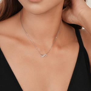 May include: A silver-toned chain necklace featuring a small infinity symbol pendant. The infinity symbol is embellished with tiny, clear, sparkling stones along one side of its loop, suspended from a delicate, thin metal link chain.