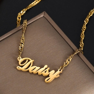 May include: A gold-coloured necklace spelling "Daisy" in a cursive font. The chain has a twisted rope design. The necklace is displayed on a brown surface against a dark background.