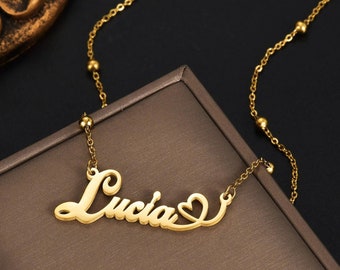 Può includere: Una collana color oro con un ciondolo nominativo che riporta la scritta Lucia in carattere corsivo, con una piccola forma a cuore integrata alla fine del nome. La catenina è a maglie sottili, arricchita da piccole perline dorate distanziate.
