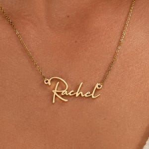 May include: A gold-toned metal necklace featuring a delicate chain and a central pendant displaying the name Rachel in a cursive script font. The nameplate is integrated directly into the chain, creating a continuous, lightweight piece of jewelry.