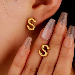 Peut inclure: Boucles d'oreilles à clous en forme de lettre "S" de couleur or. Une boucle d'oreille est dans l'oreille, et une autre est tenue dans la main. Les boucles d'oreilles sont petites et brillantes, avec un design simple et élégant.