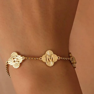 May include: A gold bracelet with a clover design. The bracelet has four clover-shaped charms, each with a letter in the center. The bracelet is worn on the wrist.