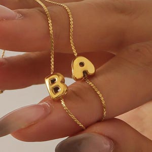 Peut inclure: Un collier doré avec les lettres "B" et "A" comme pendentifs. Les lettres sont dans une police arrondie et sont attachées à de fines chaînes en or. Le collier est tenu par une main avec de longs ongles manucurés.