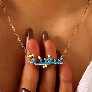 May include: A gold-colored necklace with a pendant featuring Arabic script in a turquoise color. The necklace is worn around the neck, and the chain is delicate. The pendant is the focal point, with the Arabic text clearly visible.