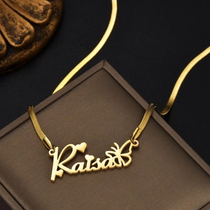 Pu&ograve; includere: Una collana color oro con un ciondolo con il nome Raisa in corsivo. Il design include due piccole forme a cuore sopra le lettere e una sagoma di farfalla attaccata sul lato destro del nome, collegata a una catenina piatta.