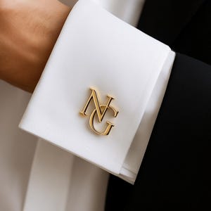 May include: Close-up of a white dress shirt cuff with a gold-colored monogram. The monogram features the letters "NG" in an elegant, stylized font. The cuff is crisp and clean, set against a black suit jacket.