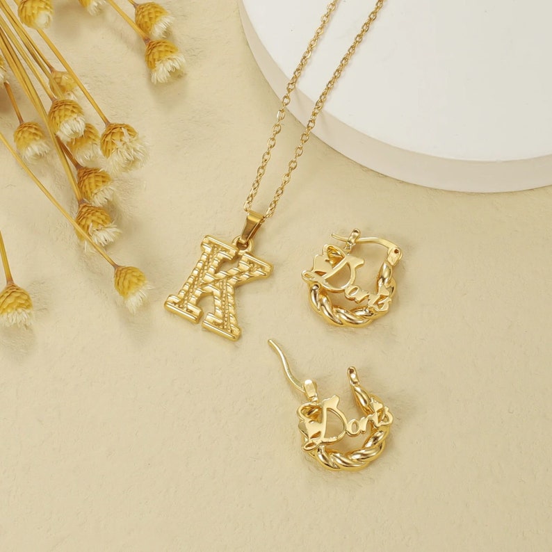 May include: Gold-toned necklace and earrings set. The necklace features a pendant with the letter K in a textured design. The earrings are hoop style with the word "Dana" in a textured design.