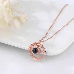 Puede incluir: Un collar de oro rosa con un colgante circular que presenta una peque&ntilde;a piedra negra y un dise&ntilde;o de hojas y peque&ntilde;as piedras blancas. El collar est&aacute; sobre una superficie blanca.