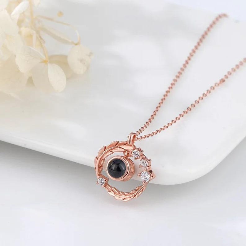 Puede incluir: Un collar de oro rosa con un colgante circular que presenta una peque&ntilde;a piedra negra y un dise&ntilde;o de hojas y peque&ntilde;as piedras blancas. El collar est&aacute; sobre una superficie blanca.