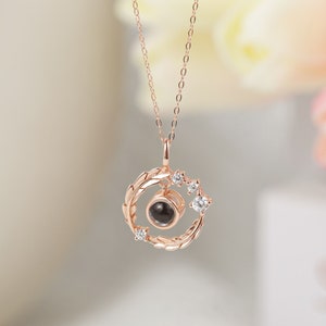 Puede incluir: Un collar de oro rosa con un colgante redondo que presenta un peque&ntilde;o c&iacute;rculo negro rodeado por un delicado dise&ntilde;o de hojas y peque&ntilde;as piedras transparentes.