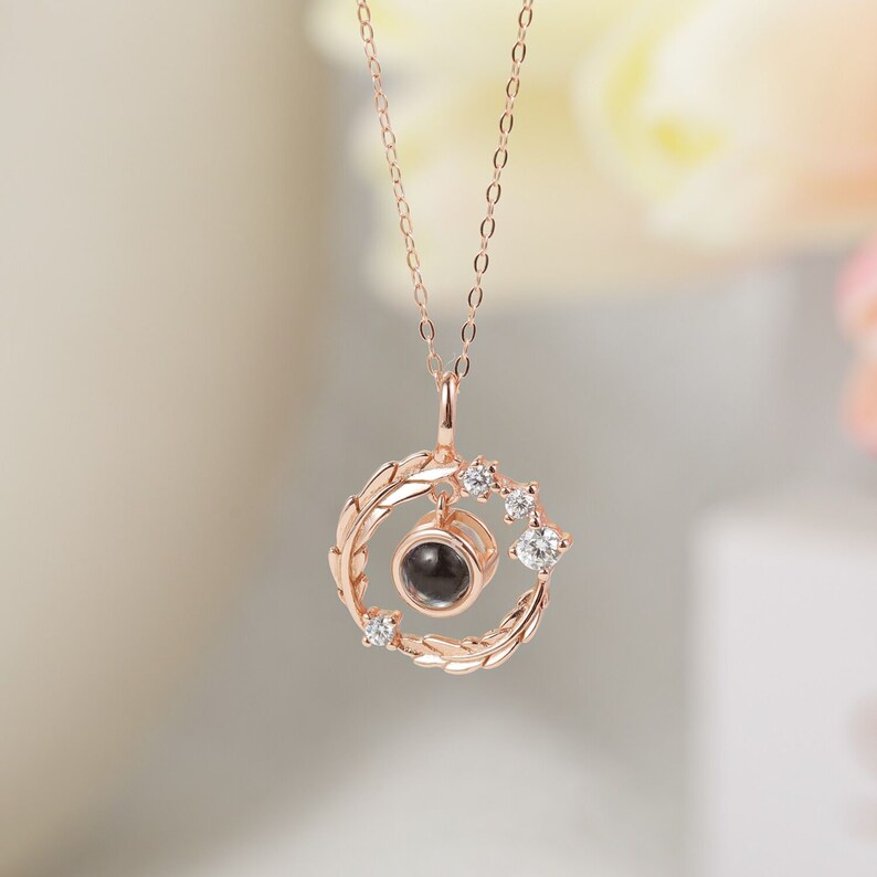 Puede incluir: Un collar de oro rosa con un colgante redondo que presenta un peque&ntilde;o c&iacute;rculo negro rodeado por un delicado dise&ntilde;o de hojas y peque&ntilde;as piedras transparentes.