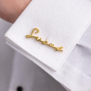 May include: Gold-colored cufflink with the name "Lucas" in cursive script. The cufflink is attached to a white dress shirt cuff. The image is a close-up, highlighting the details of the jewelry and the shirt's texture.